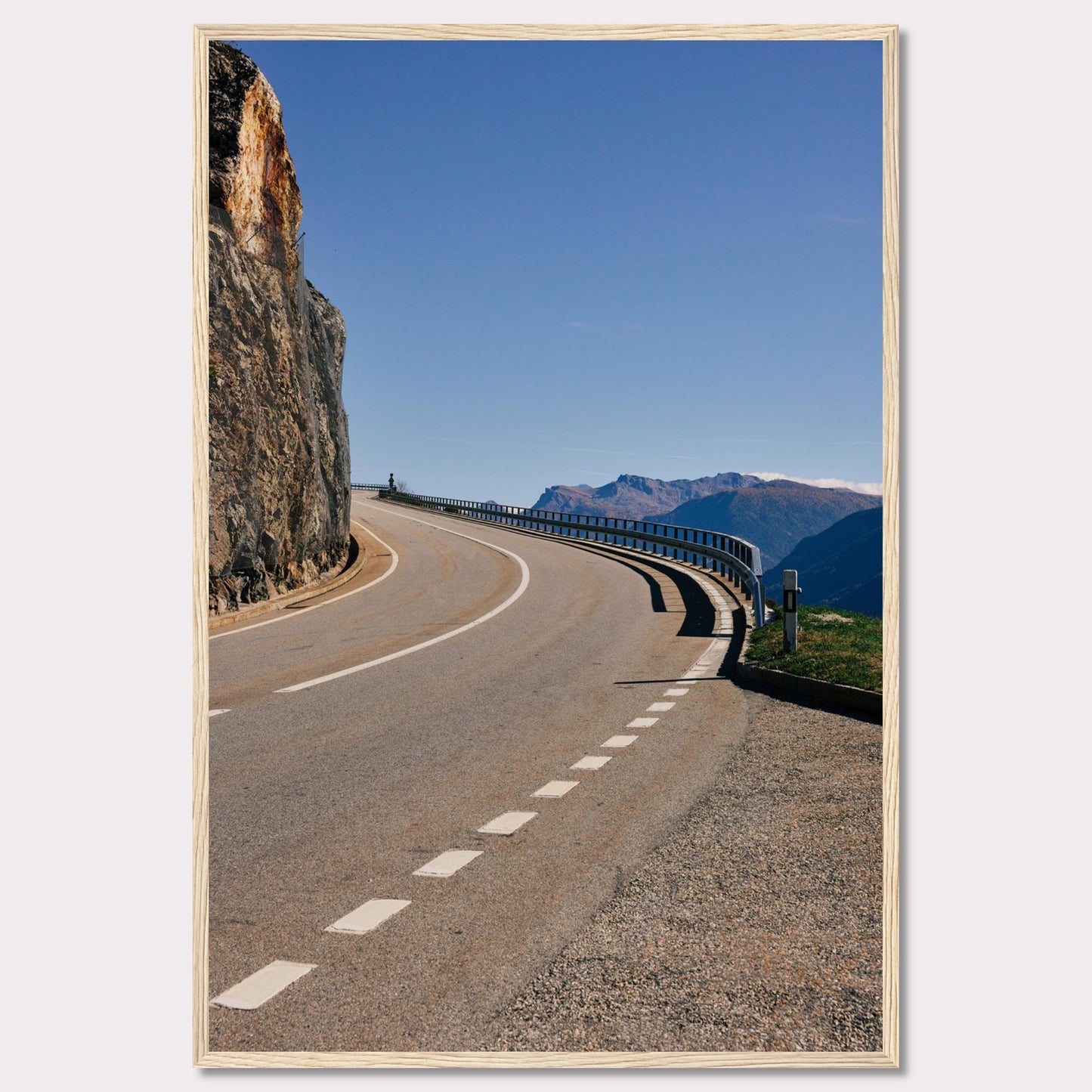 This captivating photograph showcases a winding mountain road bordered by a sheer rock face on one side and a protective guardrail on the other. The clear blue sky and distant mountain range add to the scene's serene beauty.