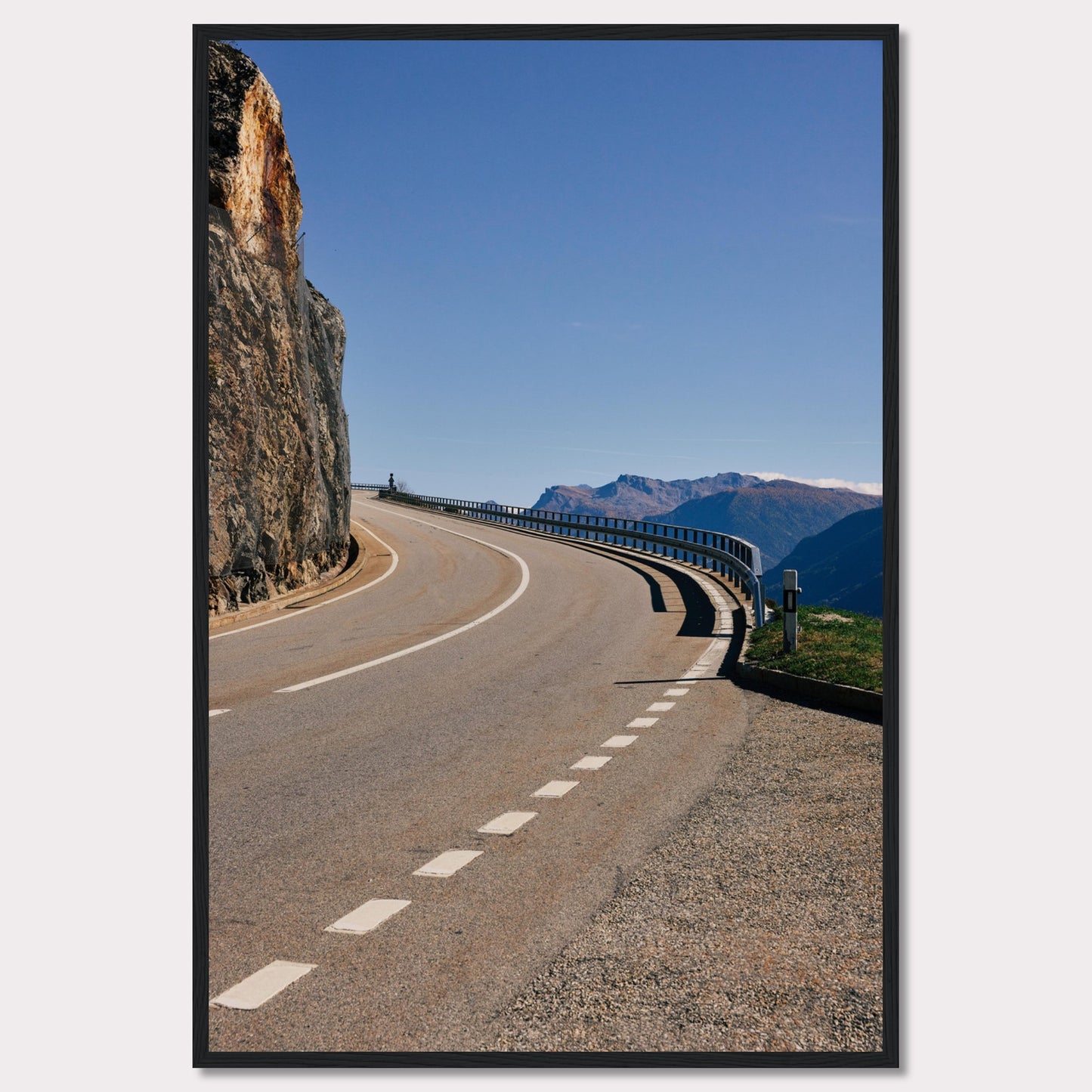 This captivating photograph showcases a winding mountain road bordered by a sheer rock face on one side and a protective guardrail on the other. The clear blue sky and distant mountain range add to the scene's serene beauty.