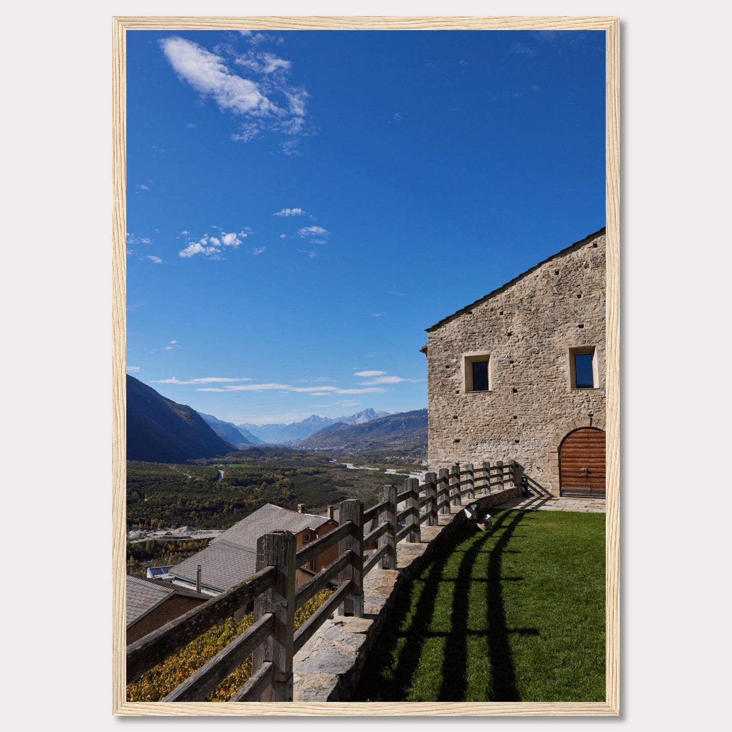 This stunning photograph captures a serene mountain landscape under a clear blue sky. The image features a rustic stone building with wooden windows and an arched wooden door. A wooden fence runs along the green grass, casting shadows that add depth to the scene. In the distance, majestic mountains stretch out, creating a breathtaking view.