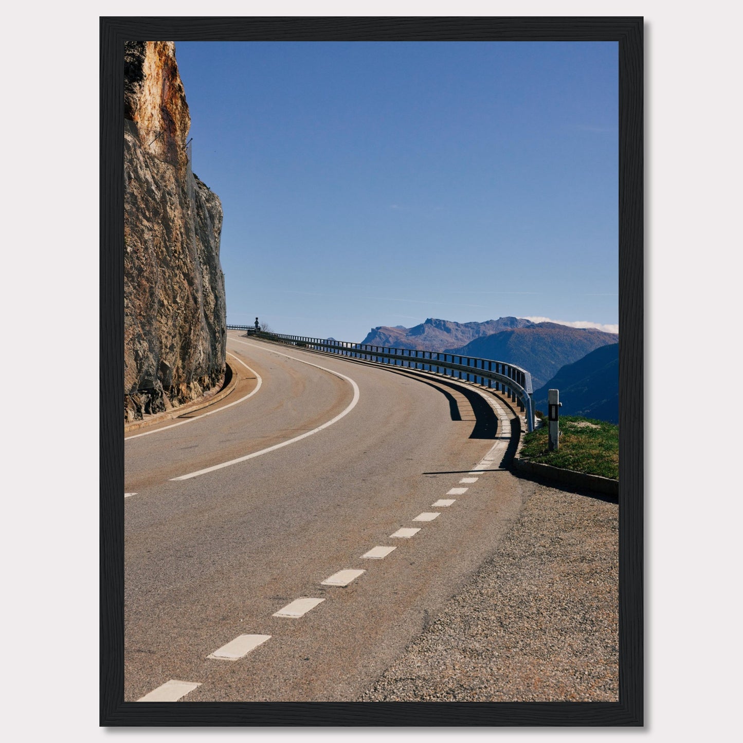 This captivating photograph showcases a winding mountain road bordered by a sheer rock face on one side and a protective guardrail on the other. The clear blue sky and distant mountain range add to the scene's serene beauty.