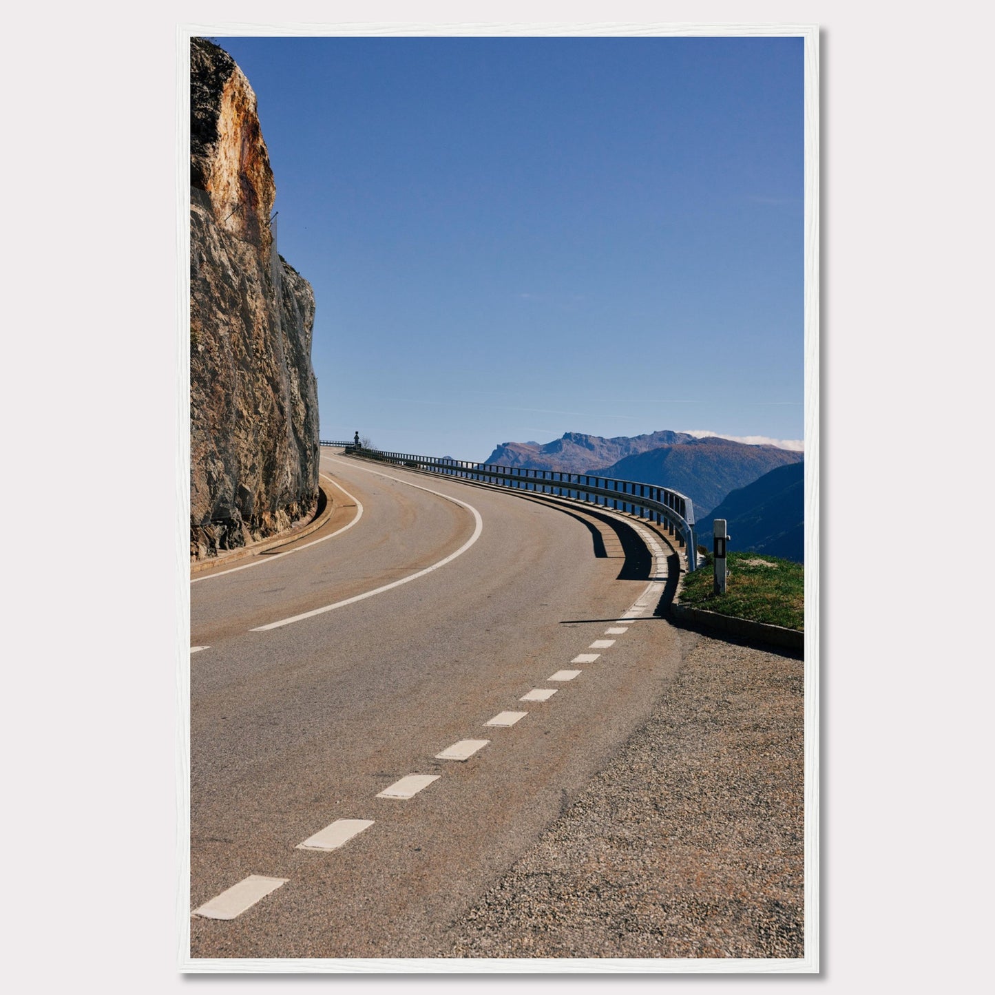 This captivating photograph showcases a winding mountain road bordered by a sheer rock face on one side and a protective guardrail on the other. The clear blue sky and distant mountain range add to the scene's serene beauty.