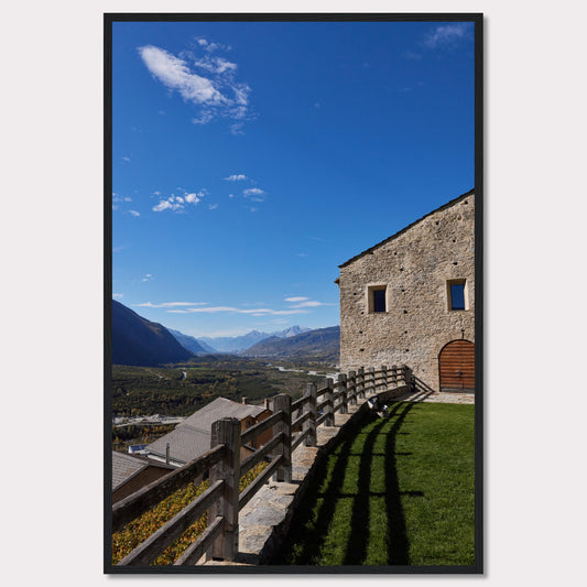 This stunning photograph captures a serene mountain landscape under a clear blue sky. The image features a rustic stone building with wooden windows and an arched wooden door. A wooden fence runs along the green grass, casting shadows that add depth to the scene. In the distance, majestic mountains stretch out, creating a breathtaking view.