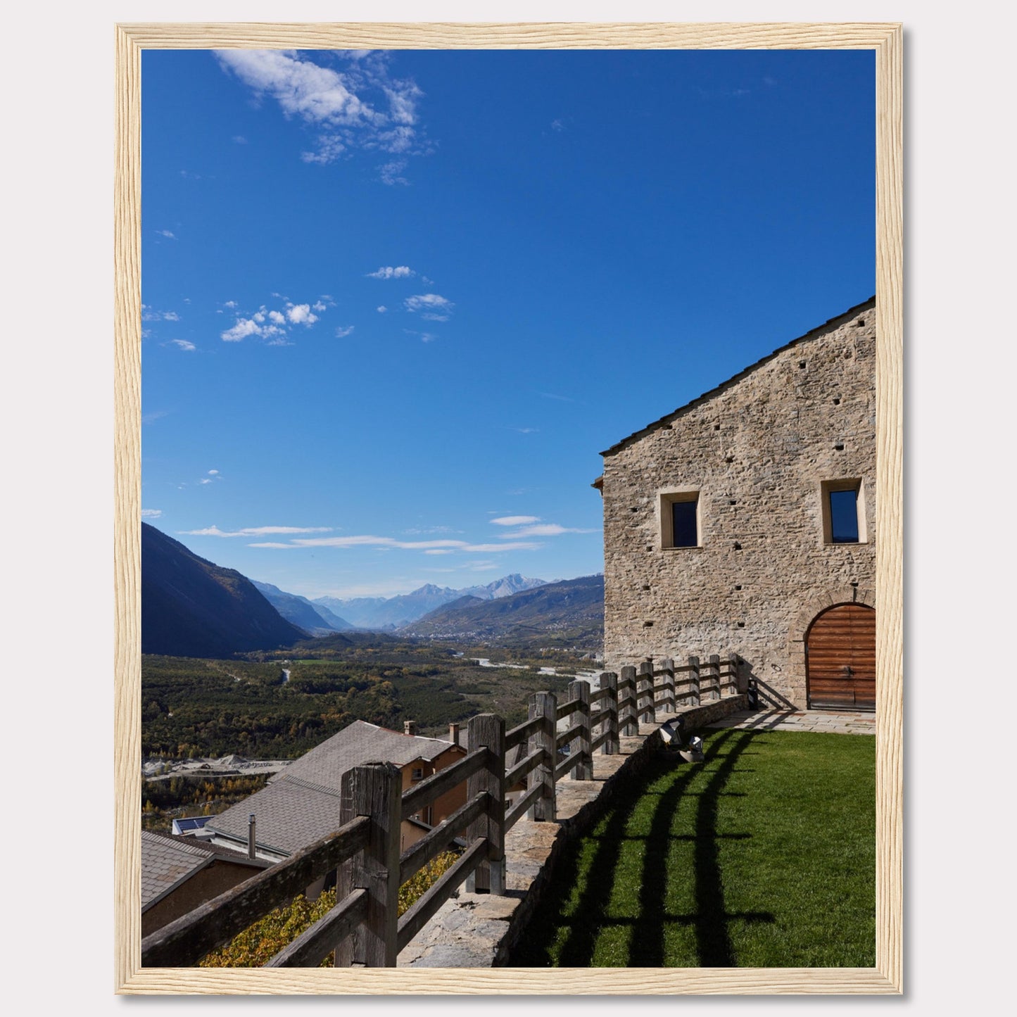 This stunning photograph captures a serene mountain landscape under a clear blue sky. The image features a rustic stone building with wooden windows and an arched wooden door. A wooden fence runs along the green grass, casting shadows that add depth to the scene. In the distance, majestic mountains stretch out, creating a breathtaking view.