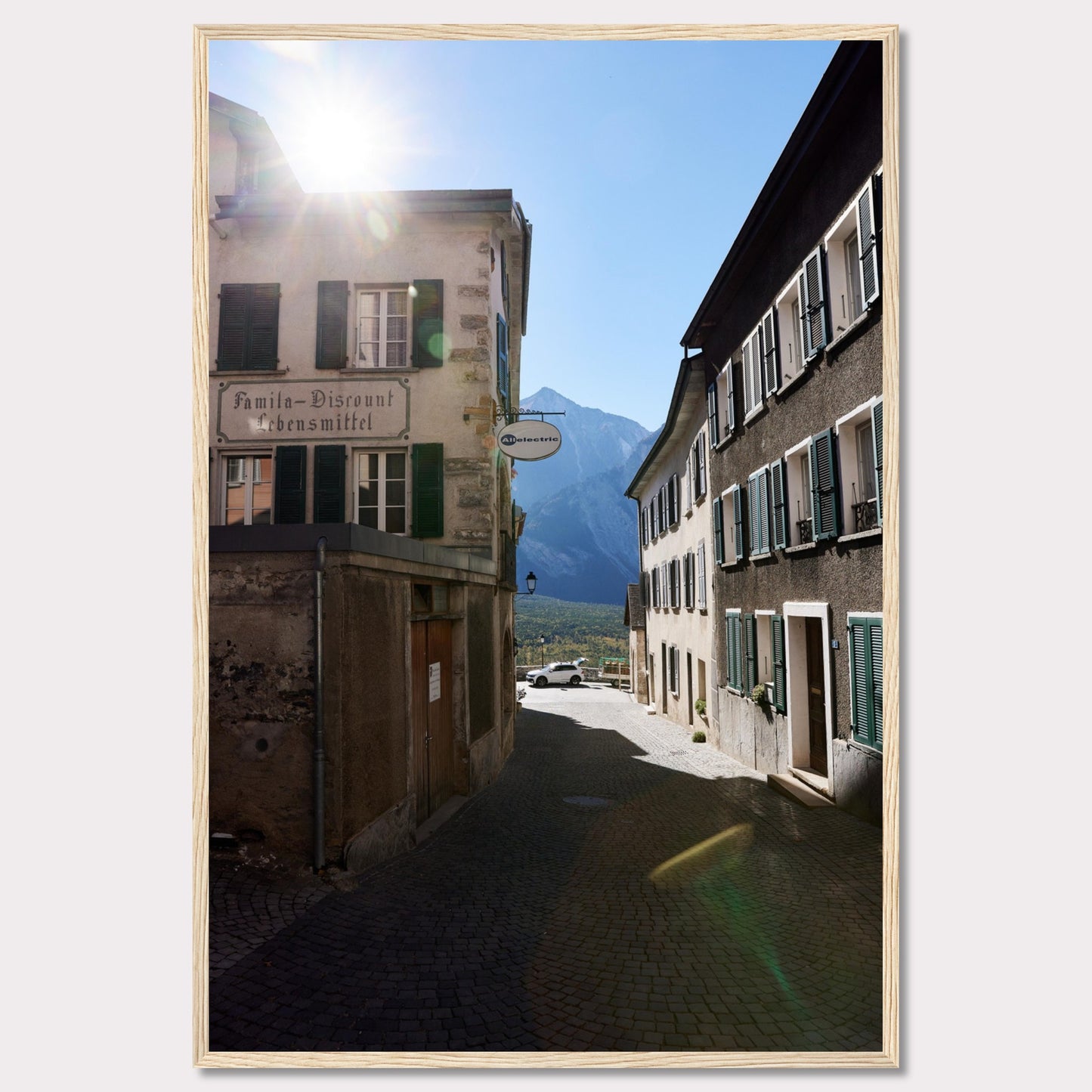 This picturesque scene captures a quaint European street bathed in sunlight, with charming buildings lining the cobblestone path. The sun peeks over the rooftops, casting a warm glow on the surroundings. In the background, majestic mountains rise against a clear blue sky.