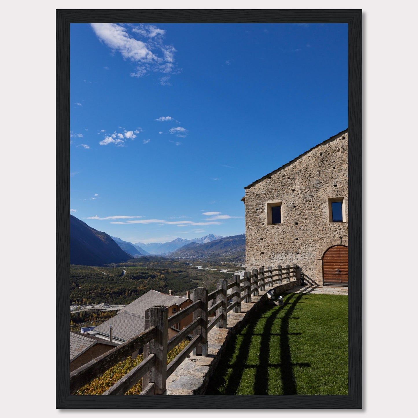 This stunning photograph captures a serene mountain landscape under a clear blue sky. The image features a rustic stone building with wooden windows and an arched wooden door. A wooden fence runs along the green grass, casting shadows that add depth to the scene. In the distance, majestic mountains stretch out, creating a breathtaking view.