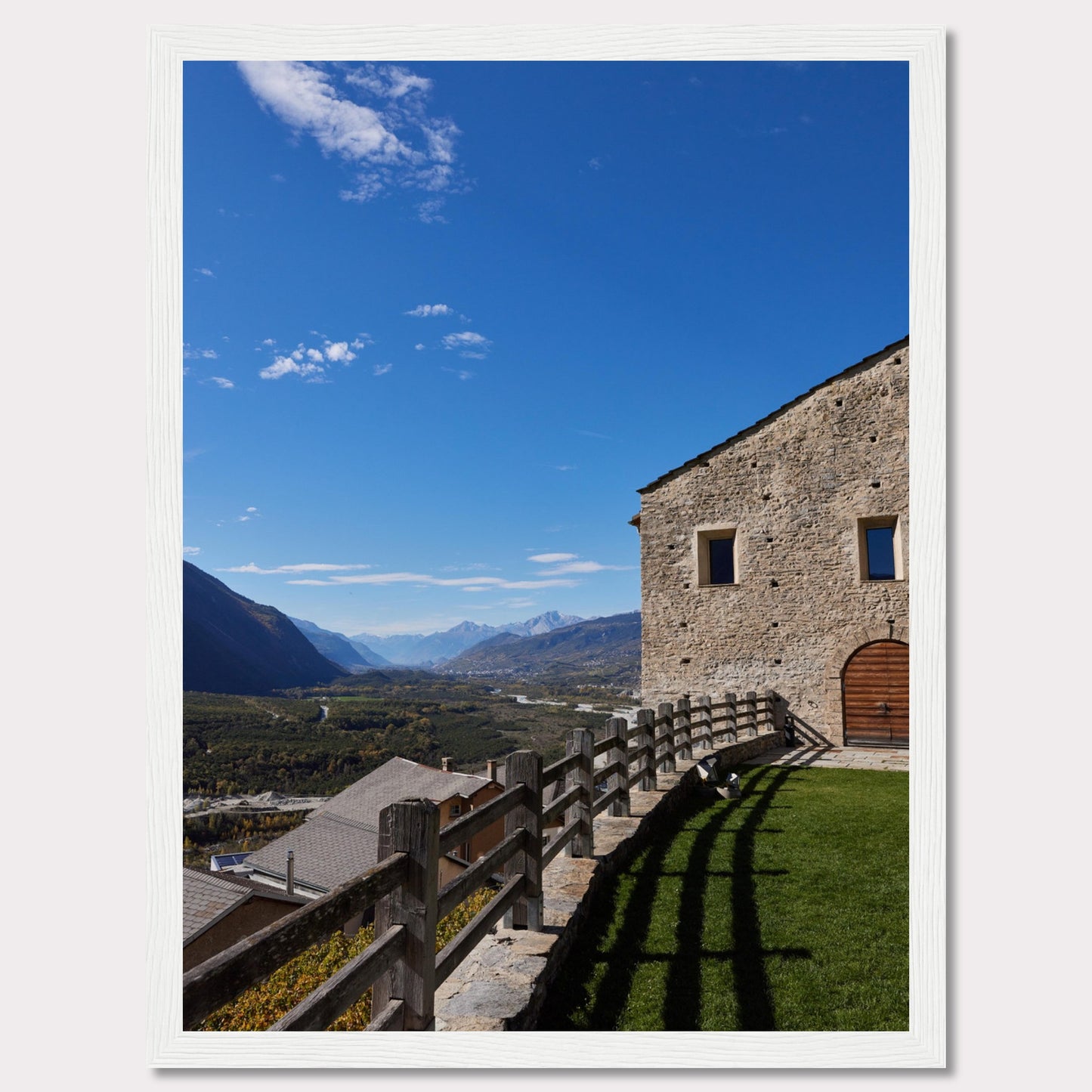 This stunning photograph captures a serene mountain landscape under a clear blue sky. The image features a rustic stone building with wooden windows and an arched wooden door. A wooden fence runs along the green grass, casting shadows that add depth to the scene. In the distance, majestic mountains stretch out, creating a breathtaking view.