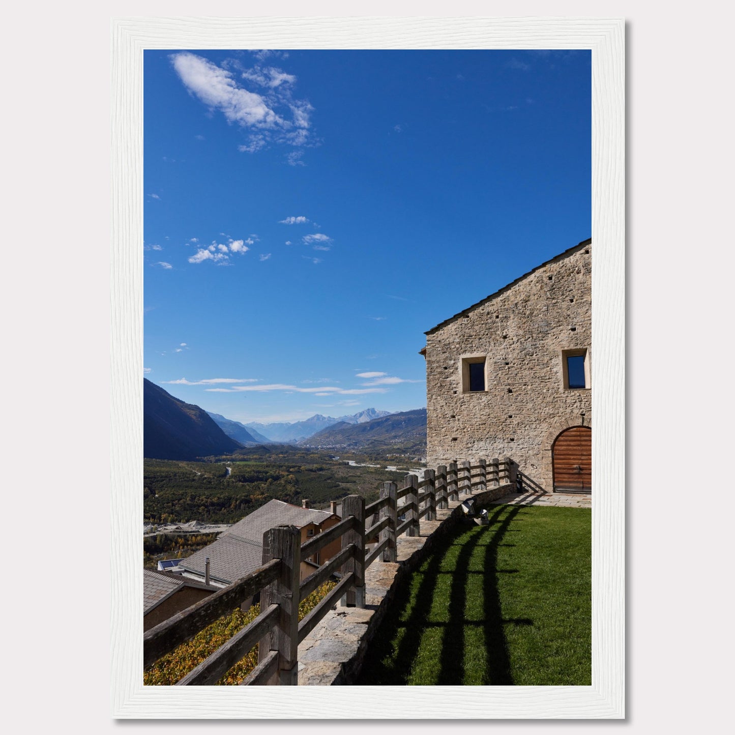 This stunning photograph captures a serene mountain landscape under a clear blue sky. The image features a rustic stone building with wooden windows and an arched wooden door. A wooden fence runs along the green grass, casting shadows that add depth to the scene. In the distance, majestic mountains stretch out, creating a breathtaking view.