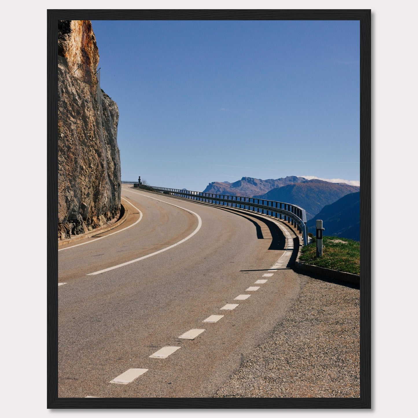 This captivating photograph showcases a winding mountain road bordered by a sheer rock face on one side and a protective guardrail on the other. The clear blue sky and distant mountain range add to the scene's serene beauty.