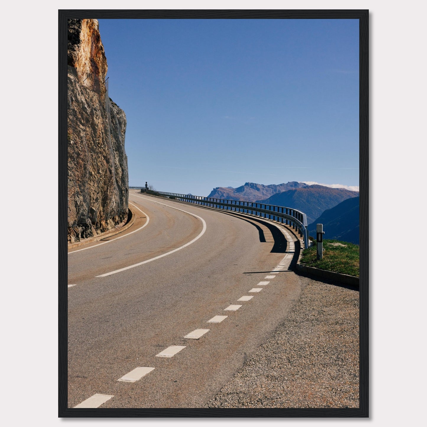 This captivating photograph showcases a winding mountain road bordered by a sheer rock face on one side and a protective guardrail on the other. The clear blue sky and distant mountain range add to the scene's serene beauty.