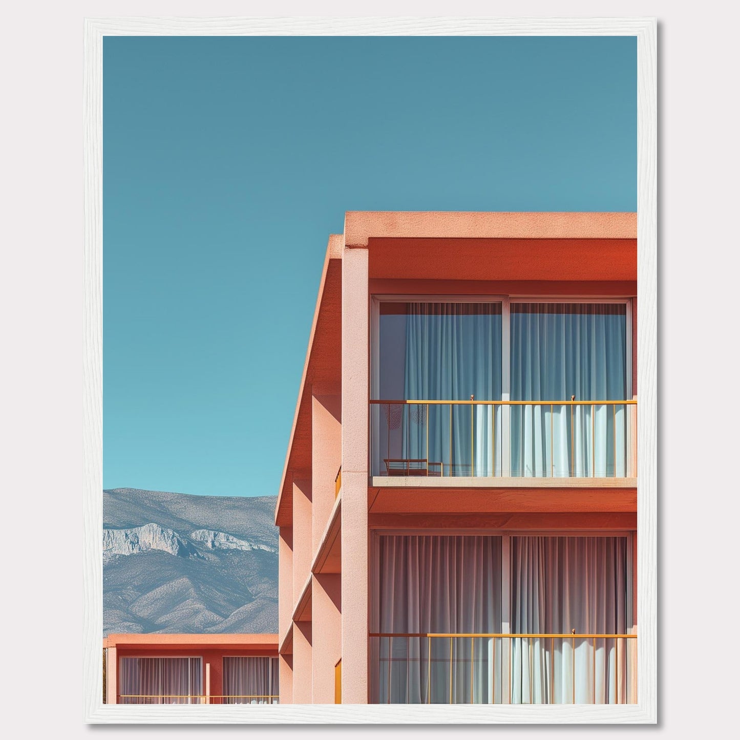 This captivating image showcases a modern building with a vibrant orange facade under a clear blue sky. The structure features large glass windows with white curtains, and balconies equipped with minimalistic furniture. In the background, majestic mountains add a serene touch to the scene.