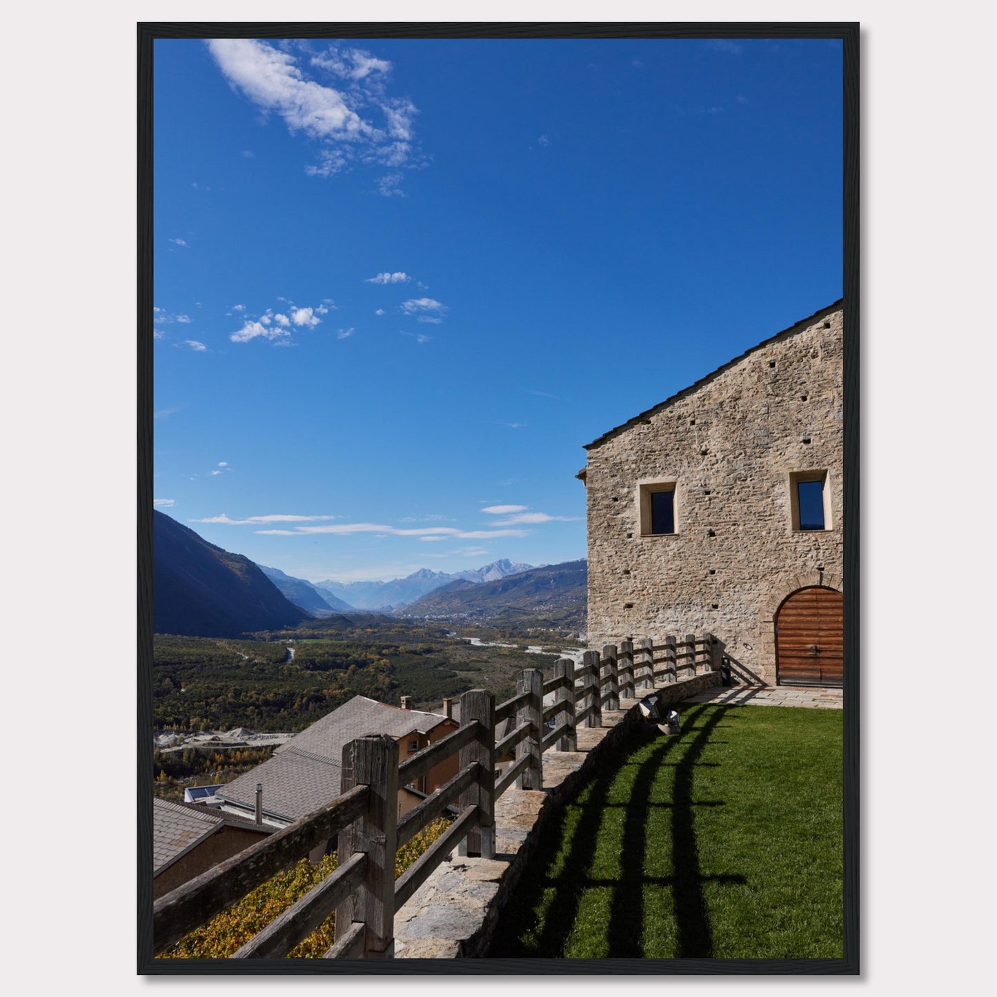 This stunning photograph captures a serene mountain landscape under a clear blue sky. The image features a rustic stone building with wooden windows and an arched wooden door. A wooden fence runs along the green grass, casting shadows that add depth to the scene. In the distance, majestic mountains stretch out, creating a breathtaking view.