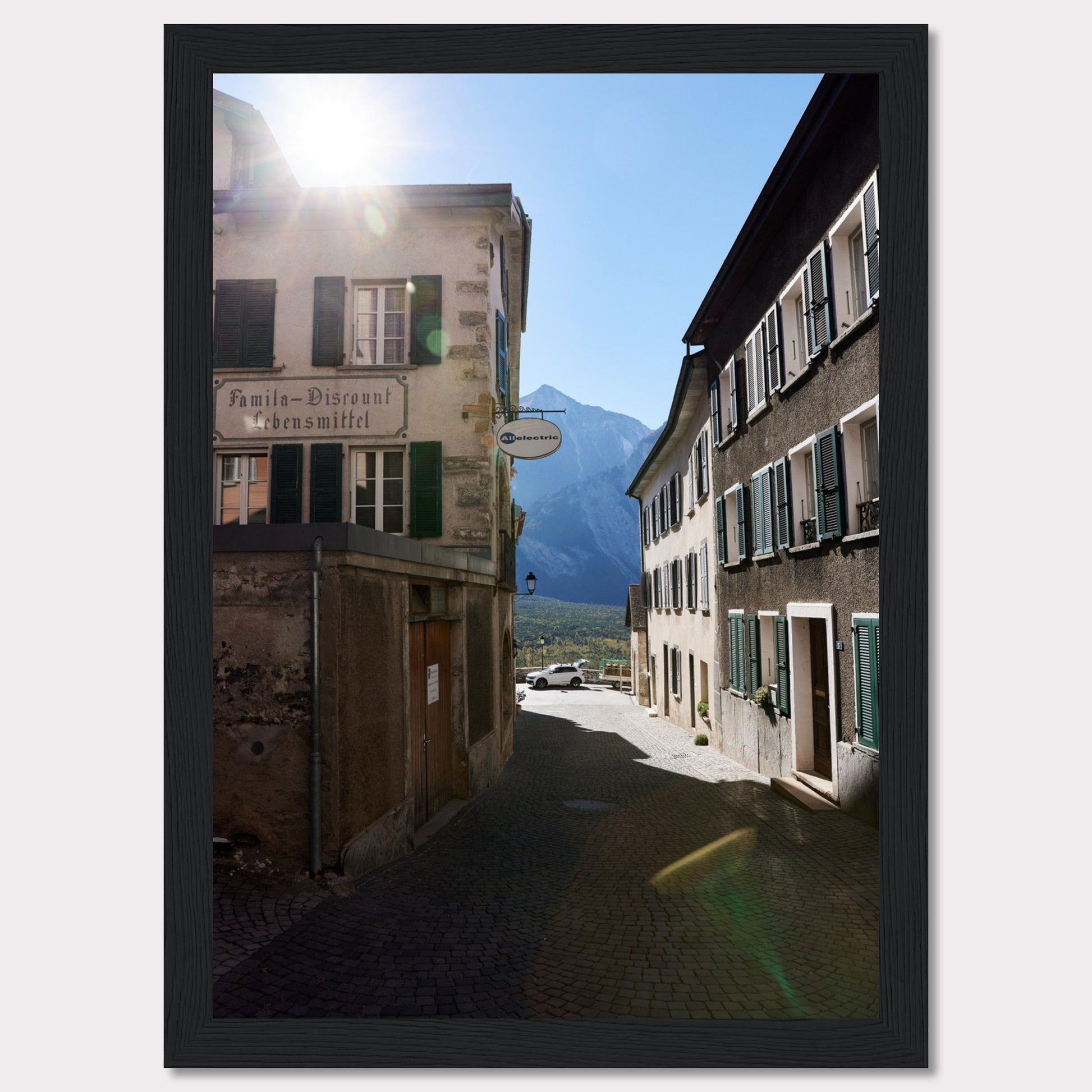 This picturesque scene captures a quaint European street bathed in sunlight, with charming buildings lining the cobblestone path. The sun peeks over the rooftops, casting a warm glow on the surroundings. In the background, majestic mountains rise against a clear blue sky.