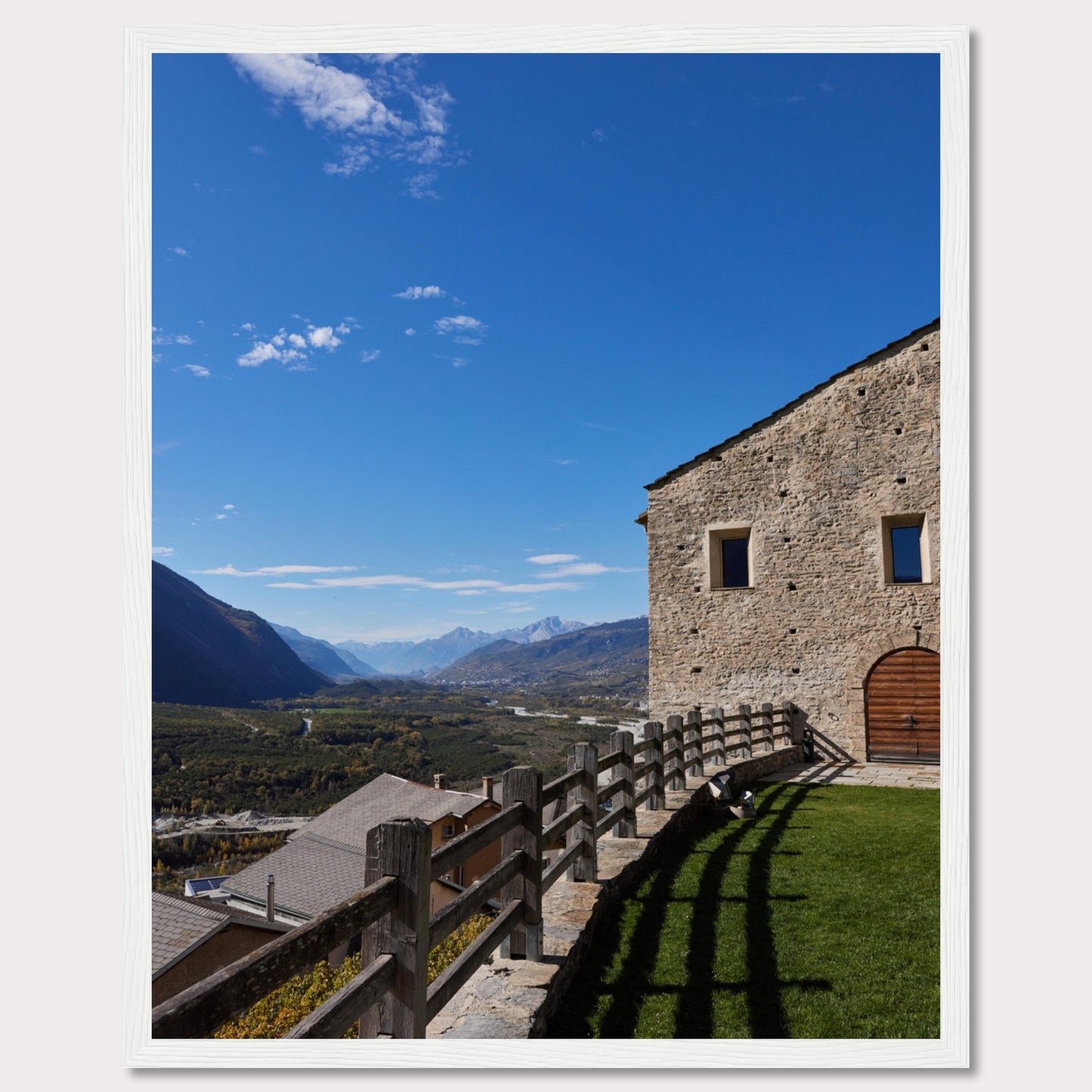 This stunning photograph captures a serene mountain landscape under a clear blue sky. The image features a rustic stone building with wooden windows and an arched wooden door. A wooden fence runs along the green grass, casting shadows that add depth to the scene. In the distance, majestic mountains stretch out, creating a breathtaking view.