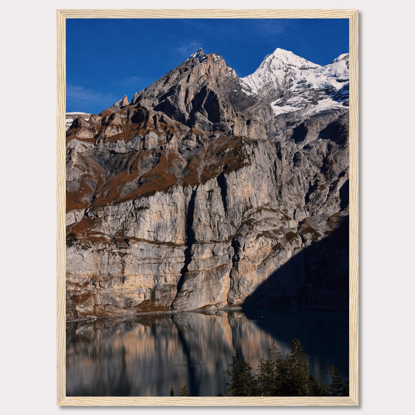 This stunning photograph captures the majestic beauty of a rocky mountain range, with snow-capped peaks under a clear blue sky. The rugged cliffs are reflected in a serene lake below, creating a mirror-like effect. Tall evergreen trees frame the bottom of the scene, adding a touch of greenery to the dramatic landscape.