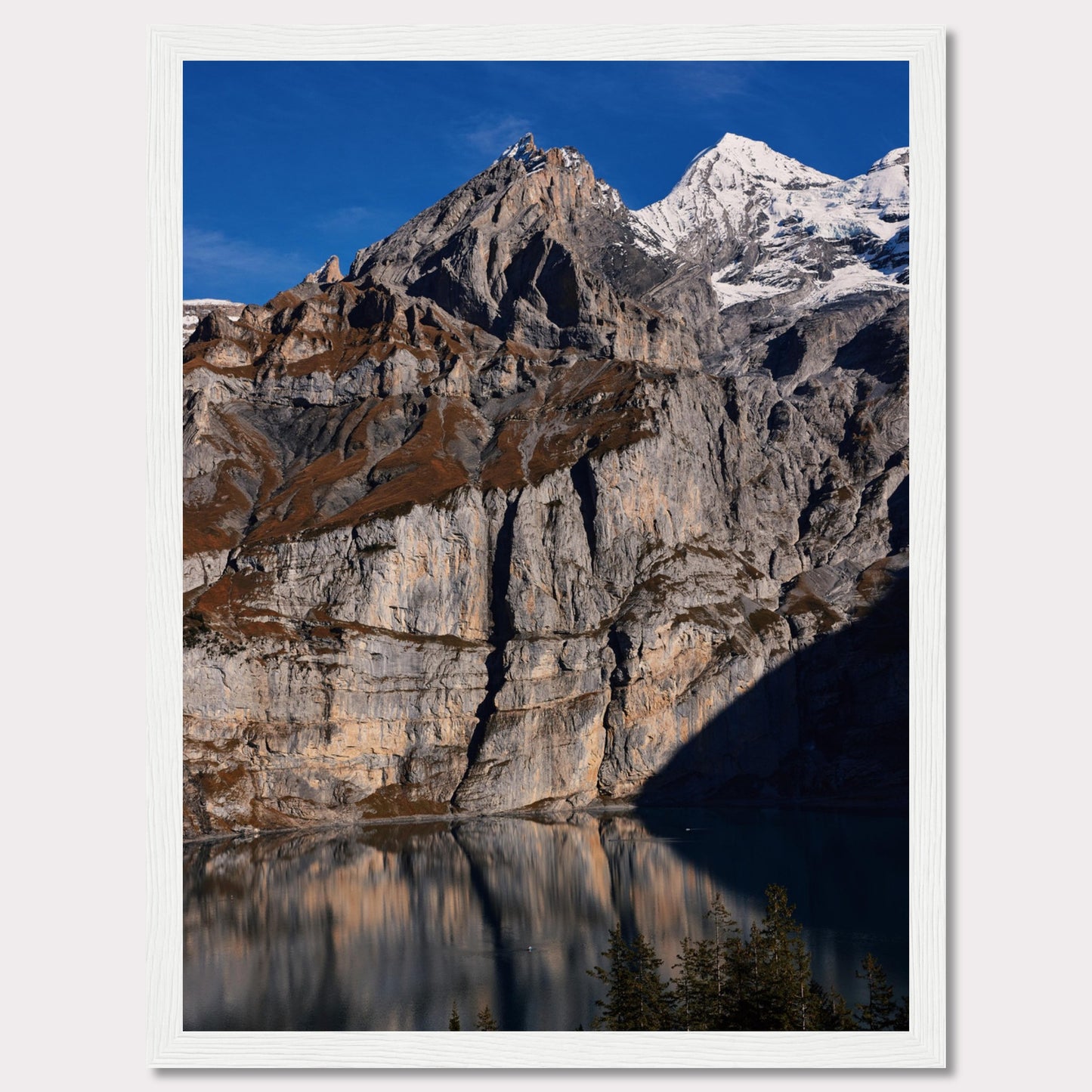 This stunning photograph captures the majestic beauty of a rocky mountain range, with snow-capped peaks under a clear blue sky. The rugged cliffs are reflected in a serene lake below, creating a mirror-like effect. Tall evergreen trees frame the bottom of the scene, adding a touch of greenery to the dramatic landscape.